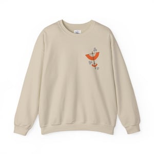 May include: Cream-colored sweatshirt with a crew neck. The design features an orange and brown abstract graphic on the left chest. The sweatshirt is made of a soft material and has long sleeves.