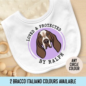 May include: White baby bib with a rounded shape. The bib features a cartoon illustration of a Basset Hound dog inside a purple circle. Text on the bib reads "LOVED & PROTECTED BY RALPH" and "ANY CIRCLE COLOUR".