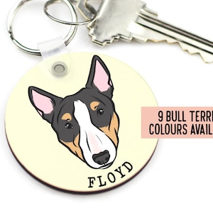Può includere: Portachiavi rotondo color crema con un'illustrazione di bull terrier in bianco e nero. Il nome del cane, "FLOYD", è stampato in nero sotto l'immagine. Il testo "9 BULL TERRIER COLOURS AVAILABLE" è stampato in nero su un rettangolo rosa.
