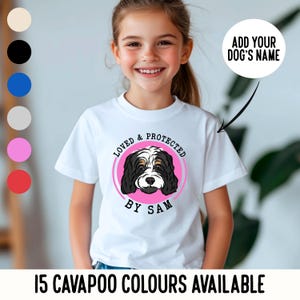 May include: White t-shirt with a pink circle featuring a black and white Cavapoo dog illustration. The text "Loved & Protected by Sam" is written around the circle.  Add your dog's name to personalise this shirt.