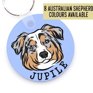 May include: A round, light blue dog tag featuring an Australian Shepherd illustration with the name "Jupile" in a curved font. The tag has a metal keyring and the text "8 AUSTRALIAN SHEPHERD COLOURS AVAILABLE" in a yellow rectangle.