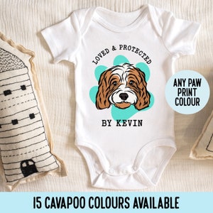 May include: White baby onesie featuring a brown and white dog illustration. The onesie reads "Loved & Protected by Kevin" and has a paw print with a turquoise background. The text "Any Paw Print Colour" is in a circle.