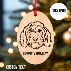 May include: A wooden oval ornament with a black and white line drawing of a Cockapoo dog. The ornament has the text "Sunny's Holiday" and "Cockapoo" printed on it.