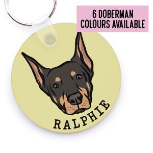 May include: Round keychain featuring a cartoon Doberman Pinscher dog head with the name "Ralphie" printed below. The background is a light yellow colour. 6 Doberman colours are available.