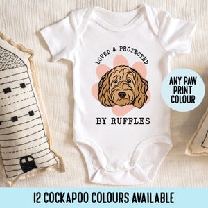 Può includere: Body bianco per neonato con un'illustrazione di un cane Cockapoo marrone e bianco. Il body dice "Loved & Protected by Ruffles" con un'impronta di zampa rosa dietro il cane. Il testo "Any Paw Print Colour" è in un cerchio blu.