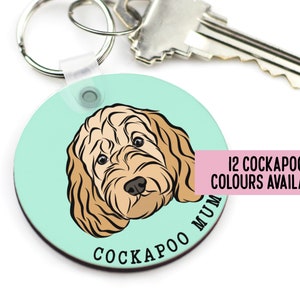 May include: Round, light blue key chain with a cartoon illustration of a Cockapoo dog. The text "Cockapoo Mum" is printed on the keychain. The text "12 Cockapoo Colours Available" is printed on a pink rectangle.