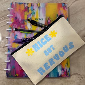 May include: A white canvas pencil case with a blue and yellow design. The text on the case reads "*NICE BUT NERVOUS*"