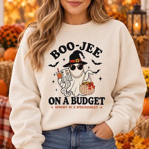 May include: A cream-colored crewneck sweatshirt featuring a graphic of a ghost wearing a witch hat, sunglasses, and a pearl necklace. The ghost holds a spreadsheet and a beaded purse. Text reads: BOO-JEE ON A BUDGET SPOOKY IN A SPREADSHEET.
