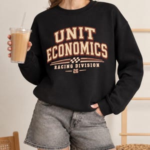 Può includere: Una felpa nera a girocollo con la scritta UNIT ECONOMICS in grandi lettere stampatello color beige e bordeaux, con le parole RACING DIVISION e il numero 26 sottostanti, accompagnata da una piccola bandiera a scacchi sul petto.