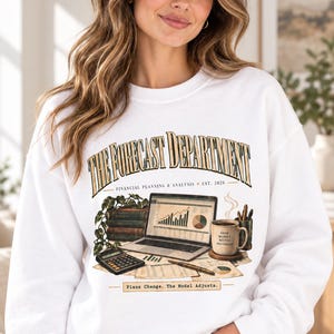 May include: A white crewneck sweatshirt featuring the text THE FORECAST DEPARTMENT, FINANCIAL PLANNING & ANALYSIS, EST. 2026, and Plans Change. The Model Adjusts. The graphic includes a laptop with charts, books, a calculator, and a coffee mug.