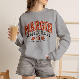 May include: A heather gray crewneck sweatshirt featuring the text MARGIN SEASON SOCIAL CLUB FINANCE DEPT in orange and black collegiate lettering, accompanied by a bar graph icon and two orange maple leaf graphics on the front chest.