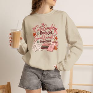 May include: A beige crewneck sweatshirt featuring a graphic print with the text "Cute Enough to Budget, Smart Enough to Forecast." The design includes illustrations of a folded sweater, a stack of books, a pink planner, a pumpkin, and autumn leaves.