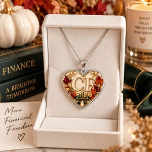Op de afbeelding: Een zilverkleurige hartvormige hanger aan een ketting met de letters CF in het midden, omringd door herfstkleurige bladeren en een kleine groene strik. De hanger wordt gepresenteerd in een wit met fluweel bekleed juwelendoosje.