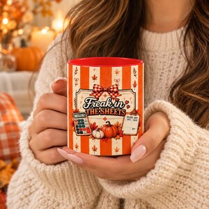May include: A ceramic mug with a red interior and an orange and white vertical striped exterior. The design features a red plaid bow, autumn leaves, a calculator, a balance sheet, and pumpkins with the text Freak in the Sheets.