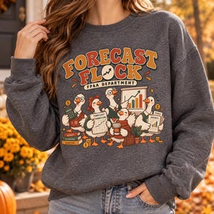 May include: A dark gray crewneck sweatshirt featuring a graphic print with the text "FORECAST FLOCK FP&A DEPARTMENT" above an illustration of five geese in business attire holding office supplies, a calculator, and financial charts.
