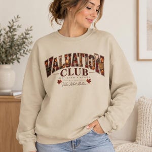 May include: A beige crewneck sweatshirt featuring the text VALUATION CLUB in a dark brown collegiate font with an autumn leaf pattern fill. Below the text are the words DCF COMPS MODELS, EST 2026, and the script text Value What Matters.