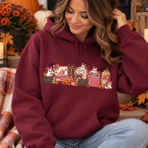 May include: A maroon hooded sweatshirt featuring a graphic print of ghosts, pumpkins, books, a computer, a calculator, and a coffee cup. The graphic includes the text "INVOICES" and "PLANS" on the books, with autumn leaves and small bows.