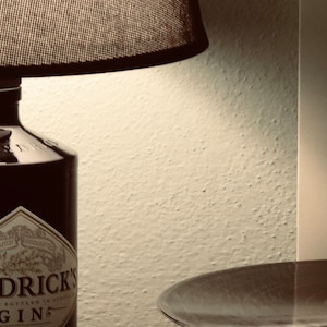 May include: A black lampshade with a black bottle of Hendricks Gin underneath. The bottle has a white label with the text "Hendricks Gin" and "Distilled and Bottled in Scotland".