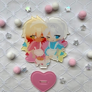 May include: An acrylic standee featuring two chibi-style characters with anime-inspired features. One character has blonde hair and yellow eyes, wearing a pink outfit, while the other has white hair and blue eyes, wearing a light blue outfit.