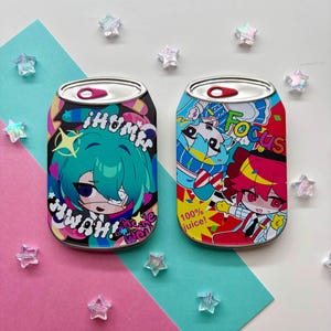 May include: Two colorful, can-shaped objects with anime-style illustrations. One features a teal-haired character with text, the other a red-haired character with the word "Focus" and "100% juice!" The cans are surrounded by star-shaped decorations.