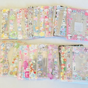May include: A collection of clear phone cases decorated with colorful cartoon stickers. The cases feature various designs, including bears, pandas, and other cute characters, with pink, blue, and yellow accents. The cases are arranged on a white surface.