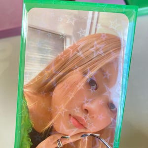 Pode incluir: Um cart&atilde;o de foto verde com uma capa de pl&aacute;stico transparente. A foto mostra uma mulher com cabelo loiro e uma camisa verde. O fundo da foto tem um padr&atilde;o de estrelas brancas.
