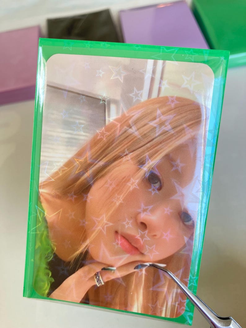 Pode incluir: Um cart&atilde;o de foto verde com uma capa de pl&aacute;stico transparente. A foto mostra uma mulher com cabelo loiro e uma camisa verde. O fundo da foto tem um padr&atilde;o de estrelas brancas.
