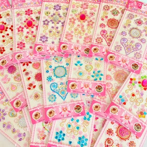 May include: A variety of colorful rhinestone stickers on pink and white packaging. The stickers feature hearts, flowers, butterflies, and other designs. The packaging says "Quick and Easy to Apply, Safe and Non Toxic".