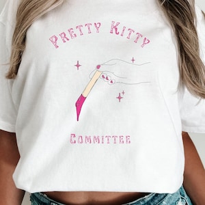 Op de afbeelding: Wit T-shirt met roze tekst die "Pretty Kitty Committee" zegt en een roze illustratie van een hand die een penseel vasthoudt met roze verf die eraf druipt.