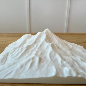 May include: A white, textured mountain sculpture with peaks and valleys. The sculpture is made of a solid material and sits on a wooden surface. The background is a white wall.