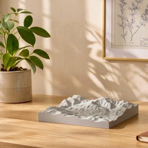 May include: A square, light grey 3D topographical map sculpture featuring detailed mountain terrain relief. The object has a flat base and textured surface representing peaks and valleys, positioned on a wooden surface next to a potted plant.