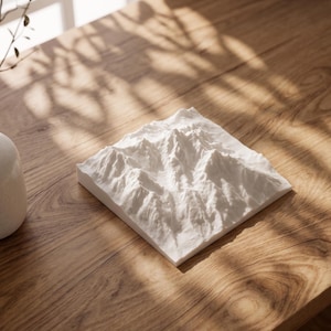 May include: A square white 3D topographic map model featuring detailed mountain peaks and valleys. The relief sculpture is crafted with a matte finish, showcasing intricate geological formations and ridges across its entire surface area.