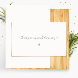Puede incluir: Una tarjeta blanca con el texto "&iexcl;Muchas gracias por tu visita!" sobre una superficie de madera con una planta verde y purpurina dorada.