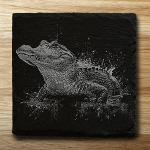 May include: Square black slate coaster with an engraved image of an alligator in water, creating a splash. The alligator is in grayscale, with detailed scales and open jaws. The coaster is on a wooden surface. The coaster measures approximately 10 cm x 10 cm.