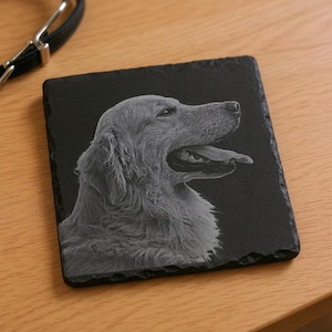 May include: A square, black slate coaster with a detailed, grayscale portrait of a Golden Retriever. The dog is shown in profile, with its mouth open and tongue out. A black leather dog collar is visible in the background.