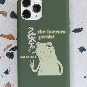 May include: Green phone case with a cartoon frog smoking a cigarette. The text on the case reads "the horrors persist but so do I".