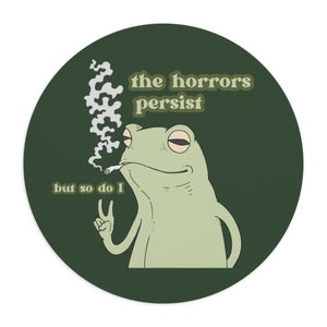May include: A green frog with a relaxed expression, holding a lit cigarette and giving the peace sign. The frog is on a dark green background with the text "the horrors persist but so do I" in a retro font.