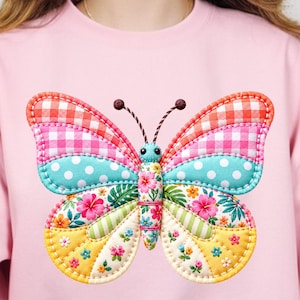 May include: Pink sweatshirt featuring a large, colorful butterfly applique. The butterfly has patchwork wings with gingham, polka dot, and floral patterns. The butterfly's body is floral, and it has brown antennae.