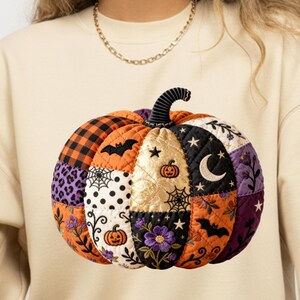 Könnte beinhalten: Ein beigefarbenes Sweatshirt mit Rundhalsausschnitt und einer großen, gesteppten Kürbisgrafik auf der Vorderseite. Der Kürbis besteht aus verschiedenen Stoffflicken in Orange, Schwarz, Lila und Gold, verziert mit gestickten Fledermäusen und Blumen.