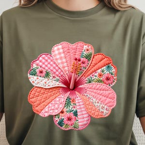 May include: Olive green t-shirt featuring a large, embroidered hibiscus flower. The flower is a patchwork design with pink, coral, and white fabrics, showcasing various patterns like gingham, polka dots, and floral prints. The flower's center has a detailed stamen.