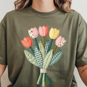 May include: An olive green t-shirt with a tulip bouquet design. The tulips are in pink, yellow, and orange, with patterned details. Green stems and leaves, tied with twine, complete the floral arrangement.
