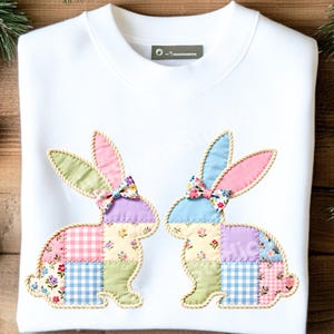 Pode incluir: Camisola branca com dois desenhos de coelhos aplicados. Cada coelho é feito de quadrados de patchwork em cores pastel, com estampas florais e xadrez. Os coelhos têm orelhas rosa e azuis e laços florais. As bordas são aparadas com um material semelhante a uma corda.