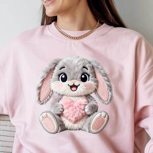 May include: A pale pink sweatshirt with an embroidered cartoon bunny holding a pink heart. The bunny has grey fur, pink inner ears, and a smiling face. The sweatshirt is a solid pale pink colour.
