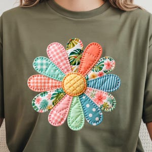 May include: Olive green t-shirt featuring a large, colorful flower applique. The flower has a yellow center and petals in various patterns, including gingham, stripes, and floral prints. The applique is stitched with a contrasting thread.