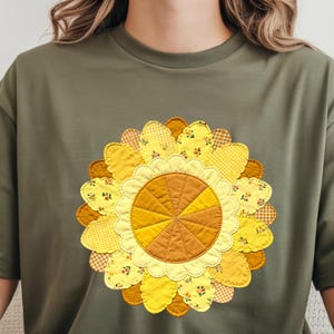 Pode incluir: T-shirt verde-oliva com um grande design de girassol aplicado. O girassol tem pétalas em camadas em tons de amarelo e castanho, com padrões florais e xadrez. Uma peça casual para o dia a dia.