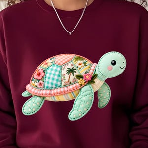 May include: A maroon sweatshirt featuring a large, quilted-style turtle graphic on the front. The turtle has a light green head and limbs with white polka dots, and a shell composed of various fabric patches including floral, palm tree, and gingham patterns.