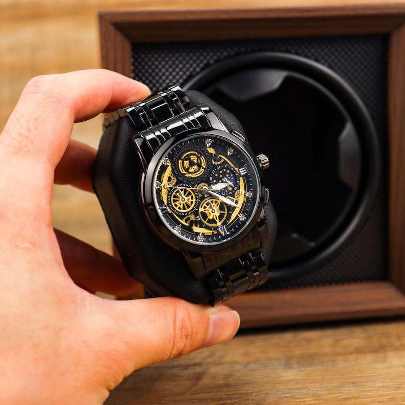 K&ouml;nnte beinhalten: Eine schwarze und goldene Skelettuhr mit einem schwarzen Metallband. Die Uhr hat ein rundes Zifferblatt mit r&ouml;mischen Ziffern und ein Datumsfenster.