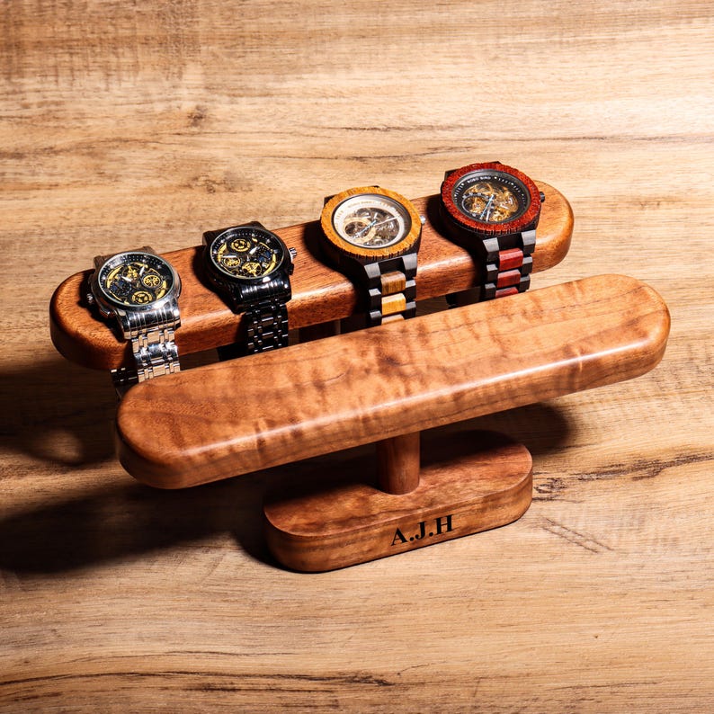 May include: Wooden watch stand with four watches on it. The stand is made of light brown wood with a dark brown stain. The watches are all different colors and styles. The stand has a small plaque on the bottom that says "A.J.H."