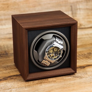 K&ouml;nnte beinhalten: Ein Uhrenbeweger aus Holz mit einem Platz f&uuml;r eine einzelne Uhr. Der Uhrenbeweger ist braun und hat ein schwarzes Innenleben. Eine silberne Uhr mit schwarzem Zifferblatt befindet sich im Uhrenbeweger.
