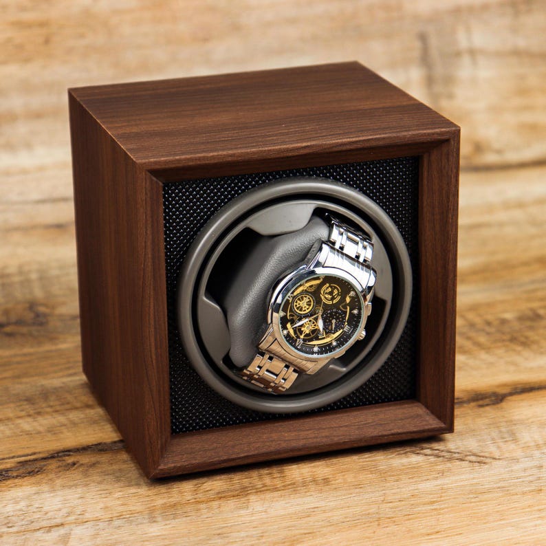 K&ouml;nnte beinhalten: Ein Uhrenbeweger aus Holz mit einem Platz f&uuml;r eine einzelne Uhr. Der Uhrenbeweger ist braun und hat ein schwarzes Innenleben. Eine silberne Uhr mit schwarzem Zifferblatt befindet sich im Uhrenbeweger.